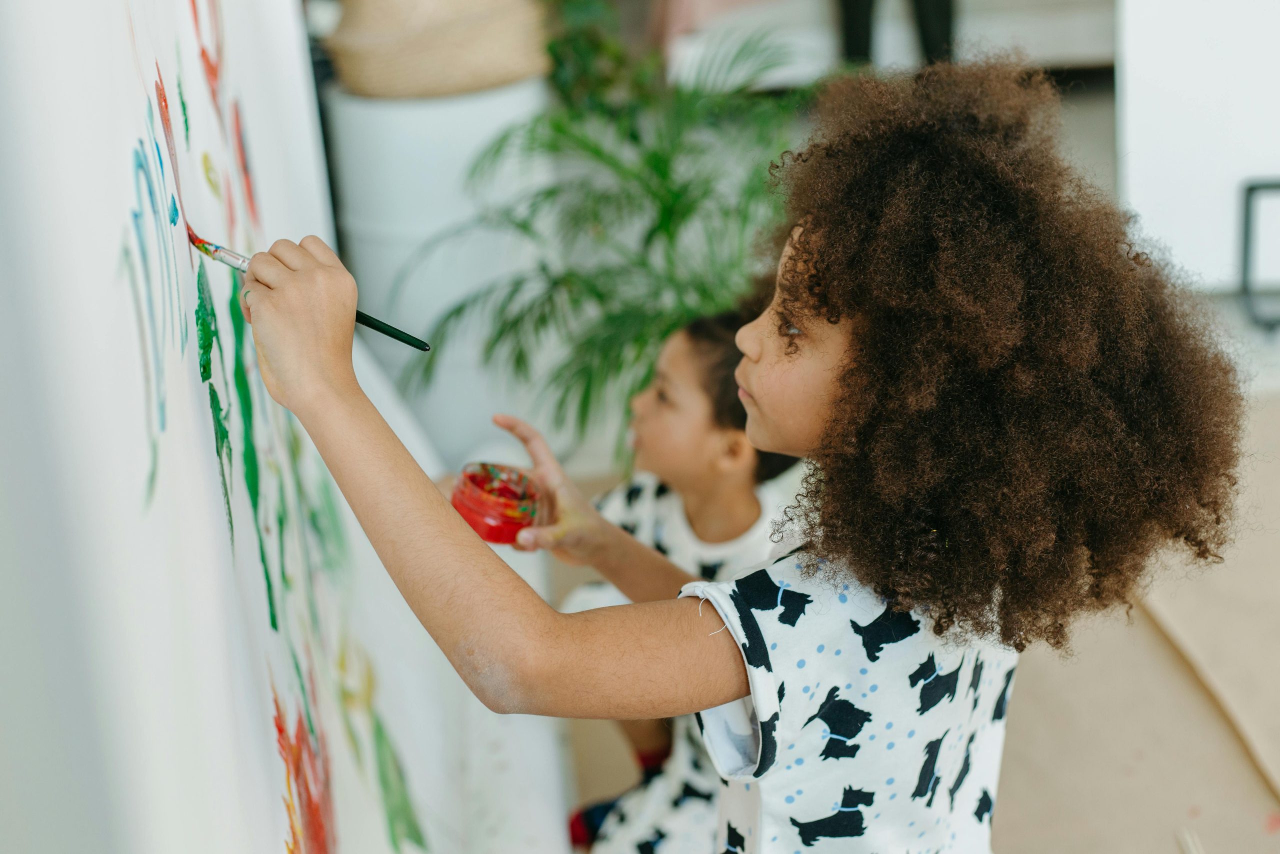Two children painting on a large white wall indoors, expressing creativity.