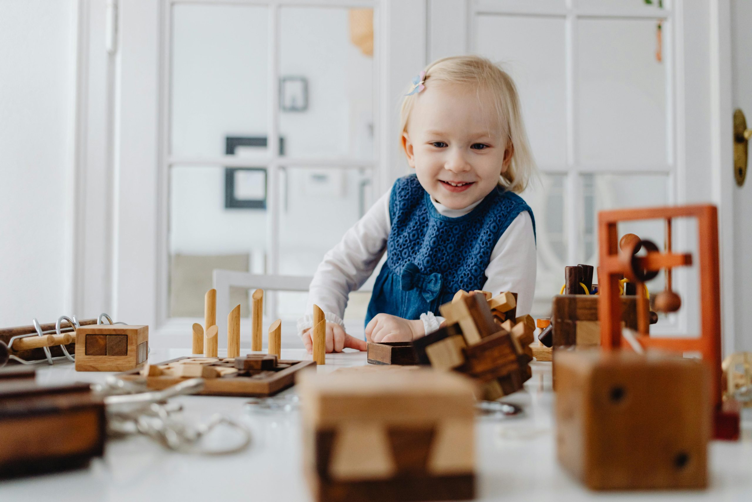 A happy toddler in a blue dress plays with wooden toys on a table indoors, enjoying a playful moment.